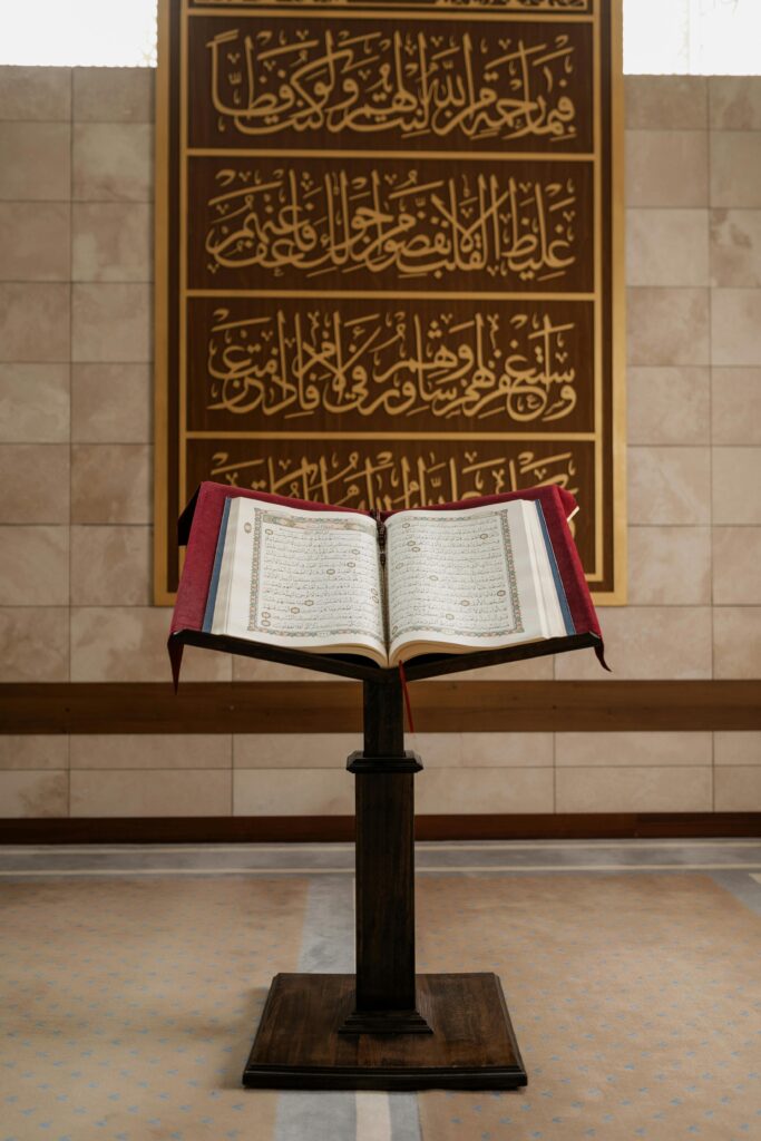 pexels-photo-8164540-8164540 A Quran open on a stand within a mosque interior, featuring Arabic calligraphy on the wall.